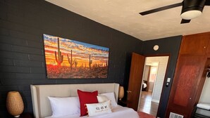 A hotel room with a bed, a ceiling fan, a painting of a desert sunset, and a door leading to another room.