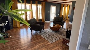 A spacious living room with wooden flooring, two armchairs, a coffee table, and a large window with curtains.