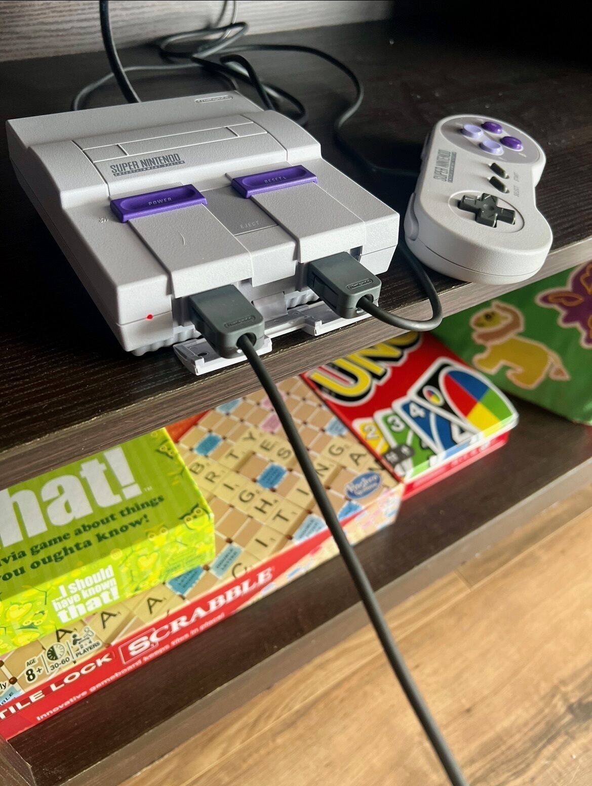 Une console Nintendo Super Nintendo branchée à une multiprise à l’aide de câbles, et une boîte de jeu de Scrabble posée sur une surface en bois.
