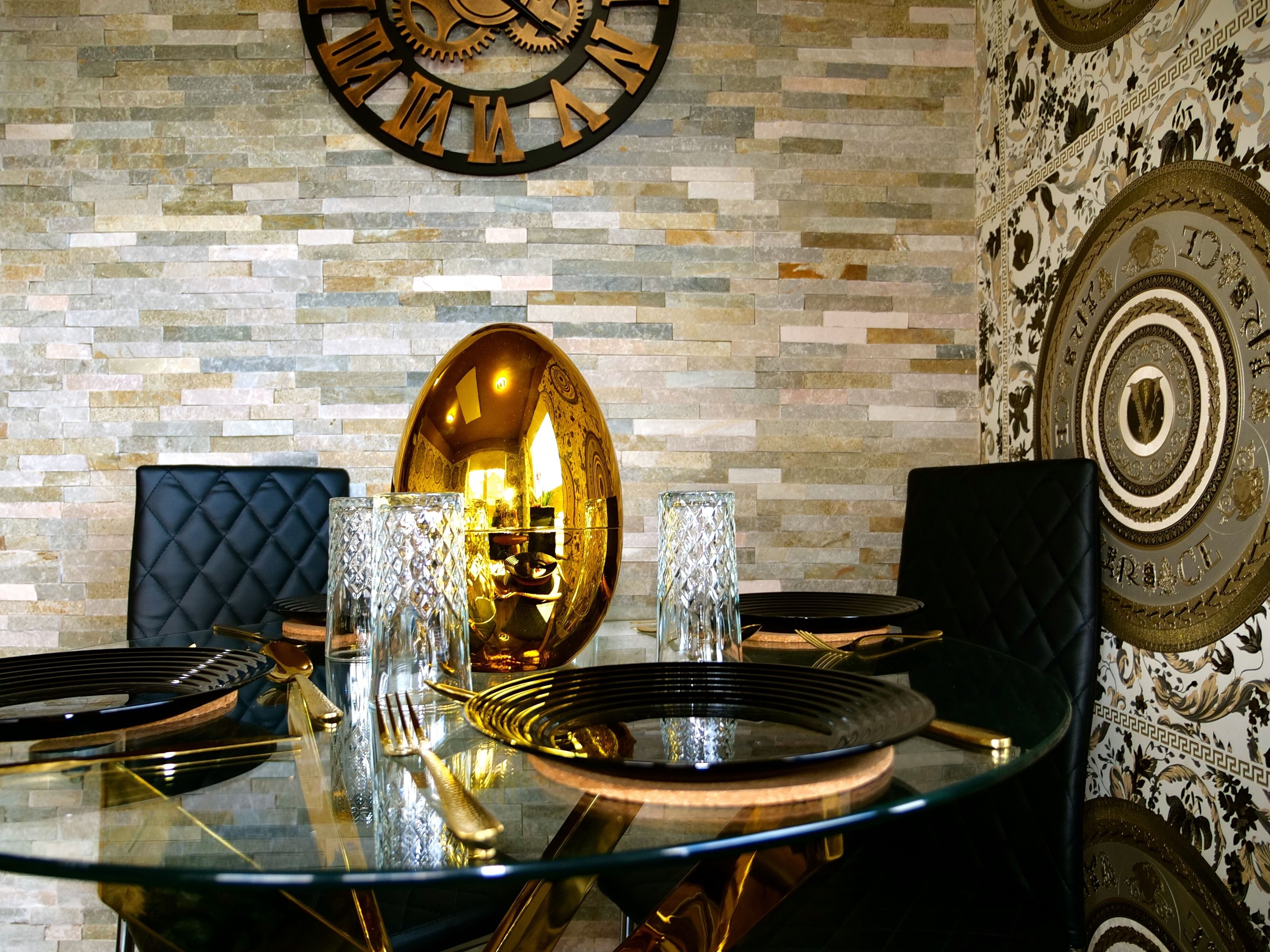 A dining table with a glass top, surrounded by black upholstered chairs, set with gold cutlery and crystal glasses, against a stone wall with a large clock and decorative plates.