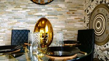 A dining table with a glass top, surrounded by black upholstered chairs, set with gold cutlery and crystal glasses, against a stone wall with a large clock and decorative plates.