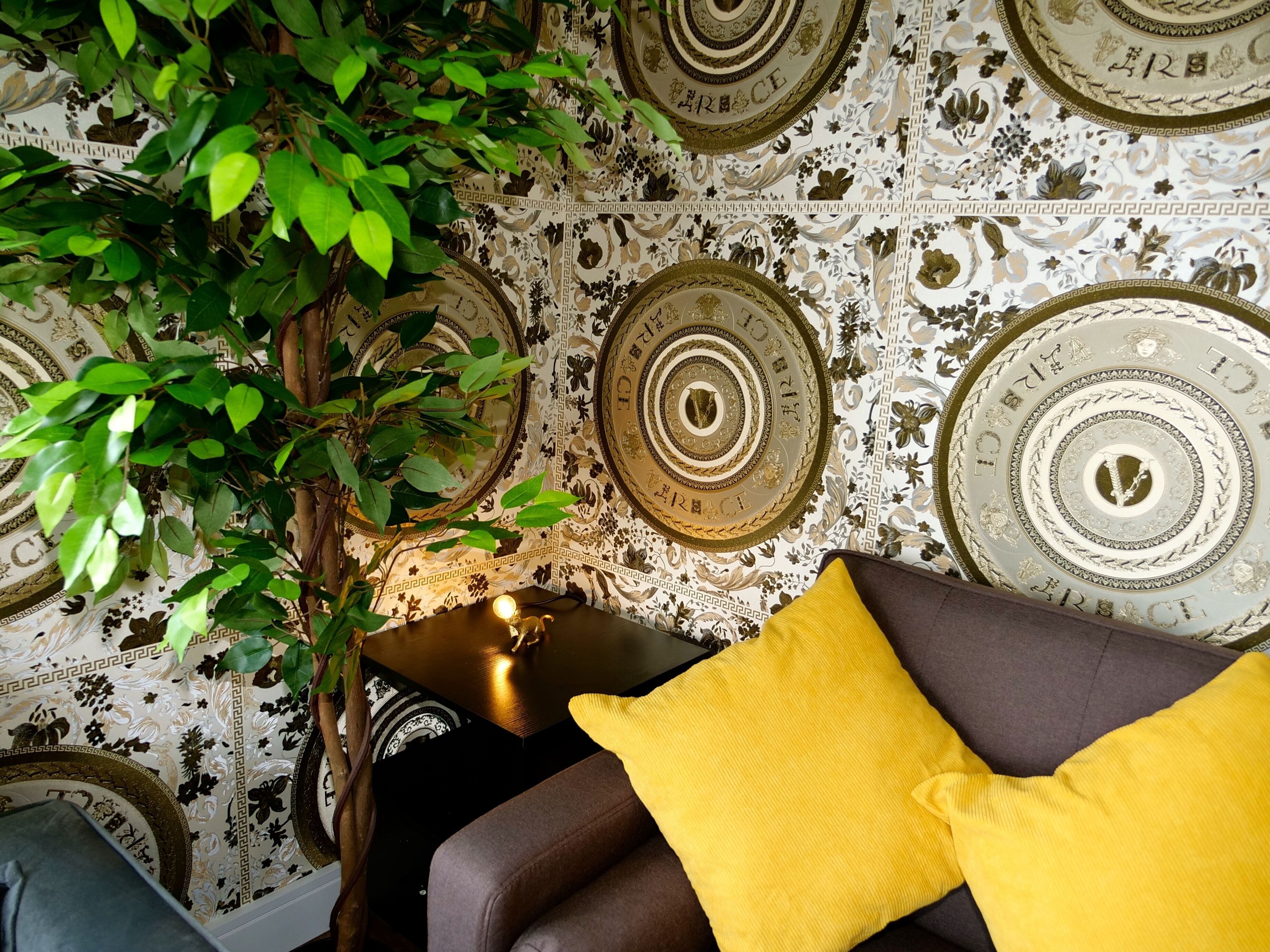 A room with a patterned wallpaper, a potted plant, a lamp, and a sofa with a yellow pillow.