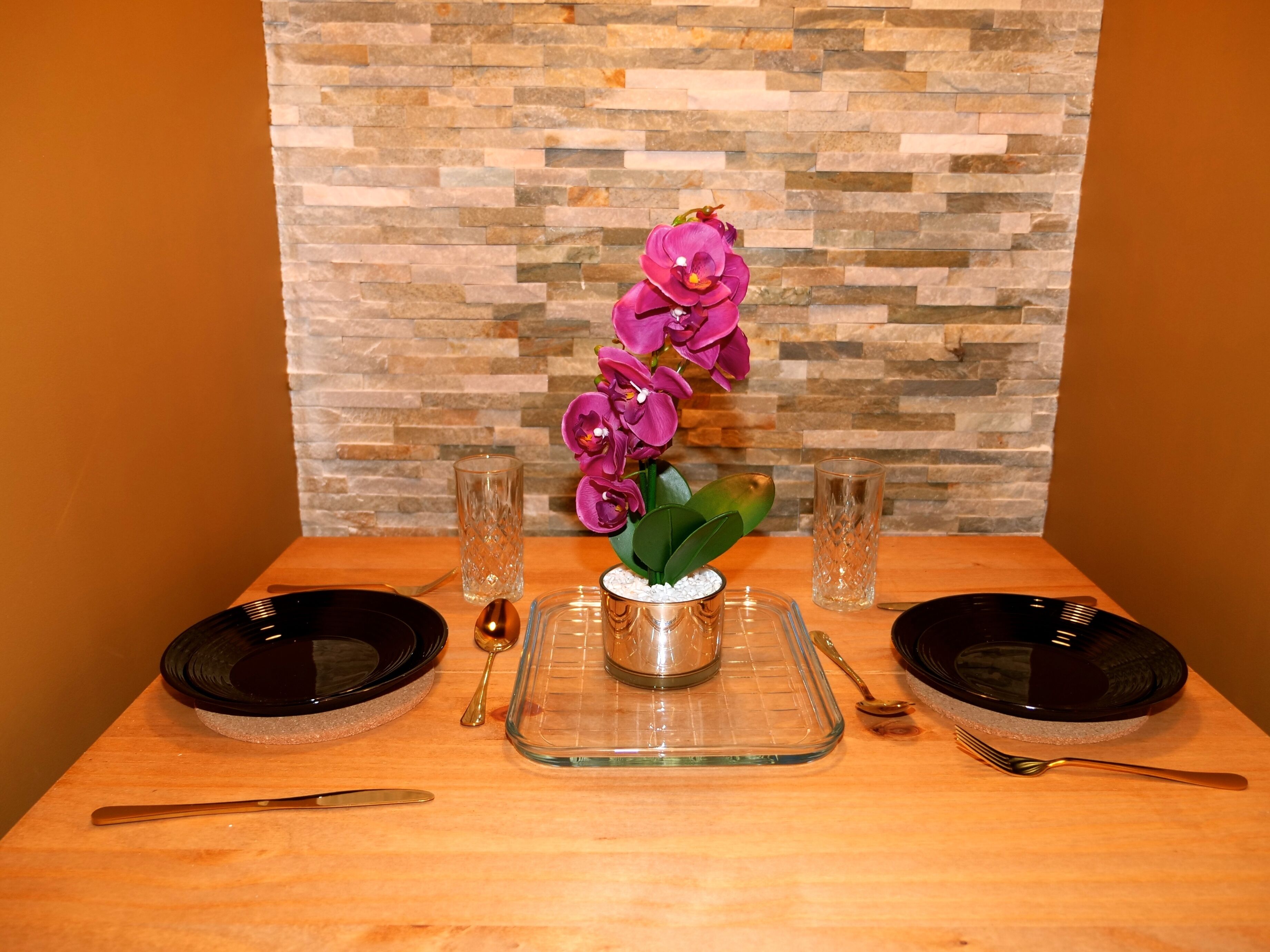 A dining table set with two black plates, a clear glass pitcher, a spoon, and a fork. A vase with purple flowers is centered on the table.