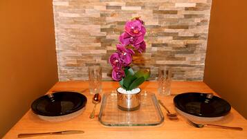 A dining table set with two black plates, a clear glass pitcher, a spoon, and a fork. A vase with purple flowers is centered on the table.