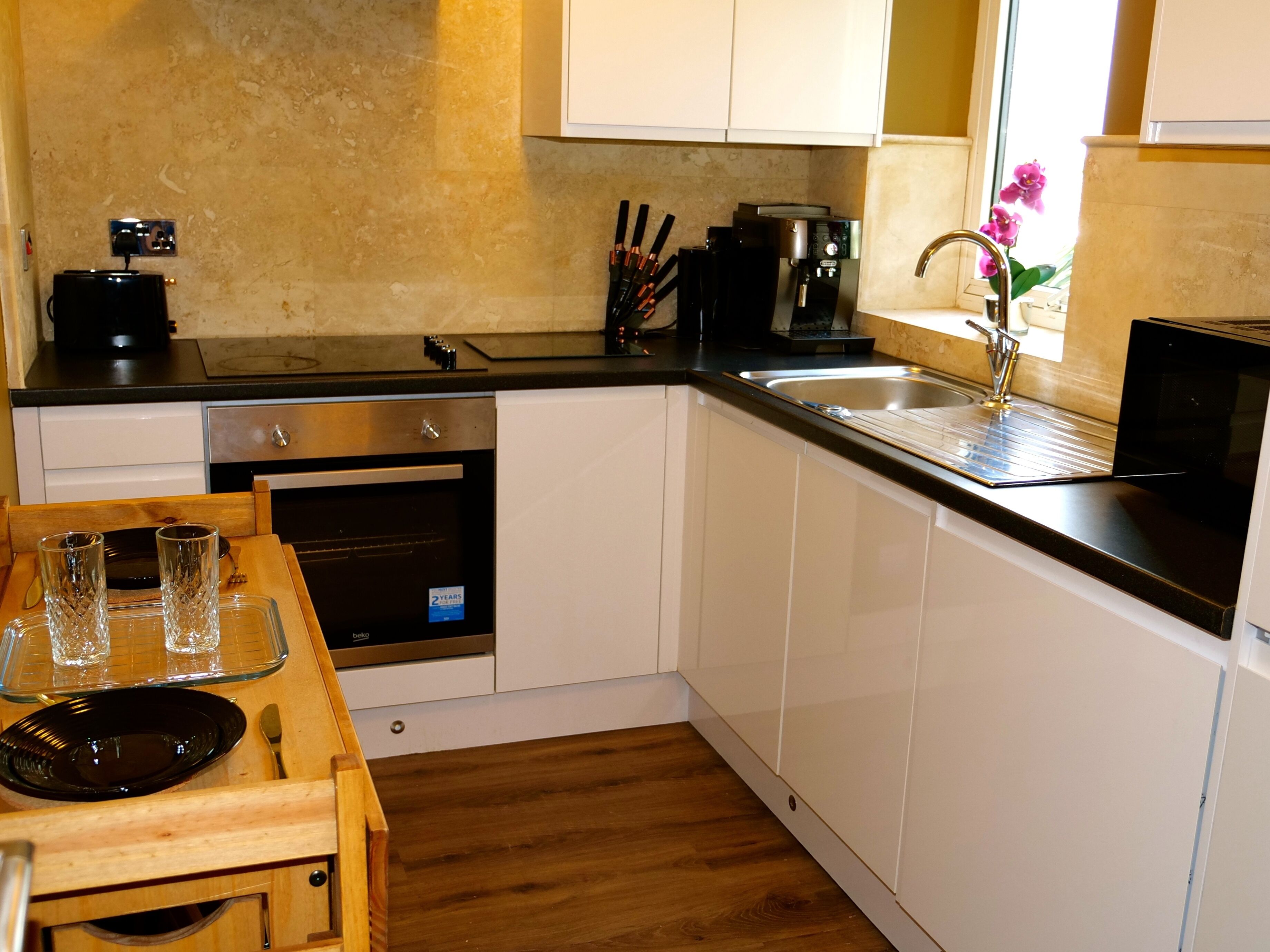 A compact kitchen with white cabinets, a black countertop, a built-in oven, and a window with a flower vase.