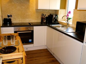 A compact kitchen with white cabinets, a black countertop, a built-in oven, and a window with a flower vase. - Deluxe Versace Styled Apartment (South Yorkshire)