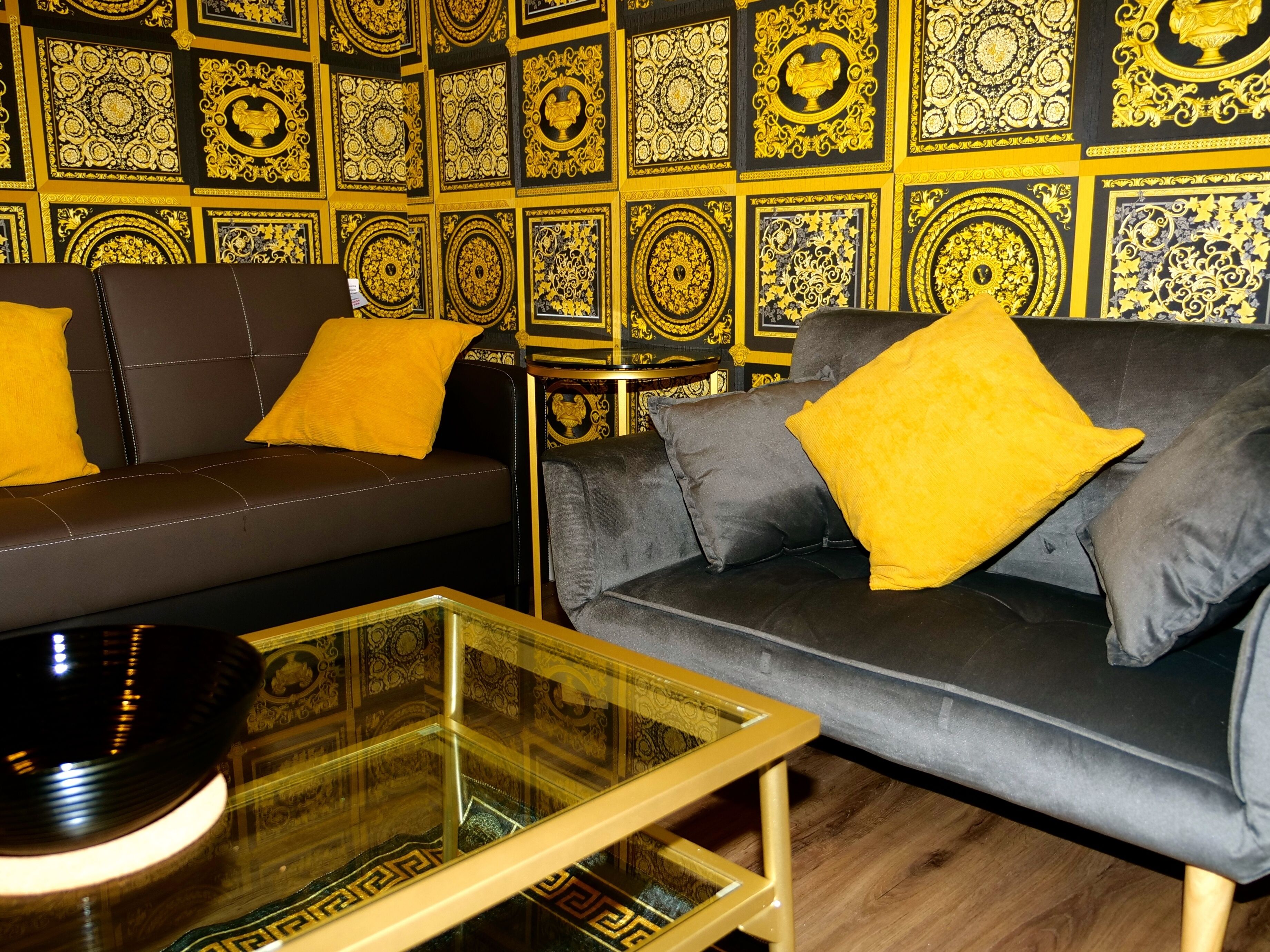 Una sala de estar moderna con un sofá marrón oscuro y un sofá gris, ambos con cojines amarillos. Una mesa de centro de vidrio está frente a los sofás. Las paredes están decoradas con intrincados diseños dorados.