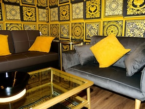 A modern living room with a dark brown sofa and a grey sofa, both with yellow cushions. A glass coffee table is in front of the sofas. The walls are decorated with intricate golden patterns.