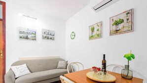 Una sala de estar con un sofá gris, una mesa de comedor de madera con una botella y un vaso, y obras de arte en la pared.