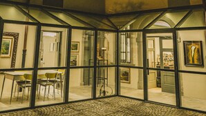Un patio con suelo adoquinado, puertas de cristal que dan a un área de comedor con sillas y mesas, y un edificio con múltiples ventanas y un balcón.