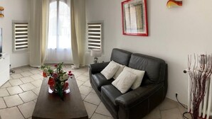 A living room with a TV on a white cabinet, a black leather sofa, a wooden coffee table with a flower vase, and a painting on the wall.