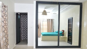 Une chambre avec une porte-fenêtre coulissante donnant sur une salle de bain, un lit recouvert d’une couvre-lit à motifs et un luminaire suspendu.