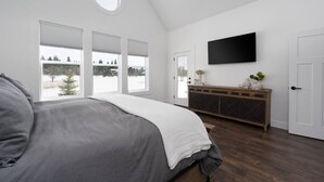 Ein Schlafzimmer mit einem großen Bett, einem an der Wand montierten Fernseher, einer Holzkommode und Blick auf Bäume.