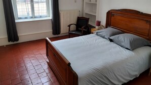 A bedroom with a wooden bed, grey pillows, a chair, a nightstand, a radiator, and a window with curtains. - Gîte de Groupes de 2 à 12 Personnes (Guillon-Terre-Plaine)