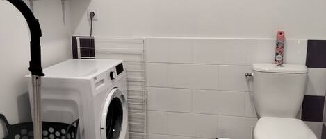 A compact laundry room with a washing machine, a drying rack, a vacuum cleaner, and a toilet.