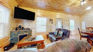 Un accogliente interno di una baita, con finiture in legno, una TV a schermo piatto montata a parete, un divano in pelle, un tavolino da caffè e un tavolo da pranzo con sedie.