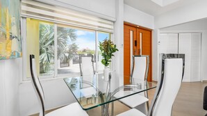 Un espace repas moderne avec une table en verre et des chaises blanches, de grandes fenêtres équipées de stores et une vue sur des palmiers.