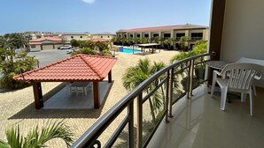 Un balcón con vista a una piscina, un espacio de estar cubierto y un edificio con tejado rojo.