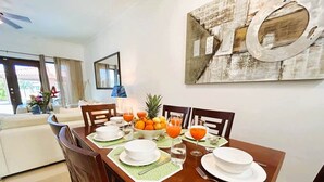 Un comedor con una mesa de madera dispuesta para cuatro personas, rodeada de sillas. Hay un frutero y vasos de jugo de naranja sobre la mesa.