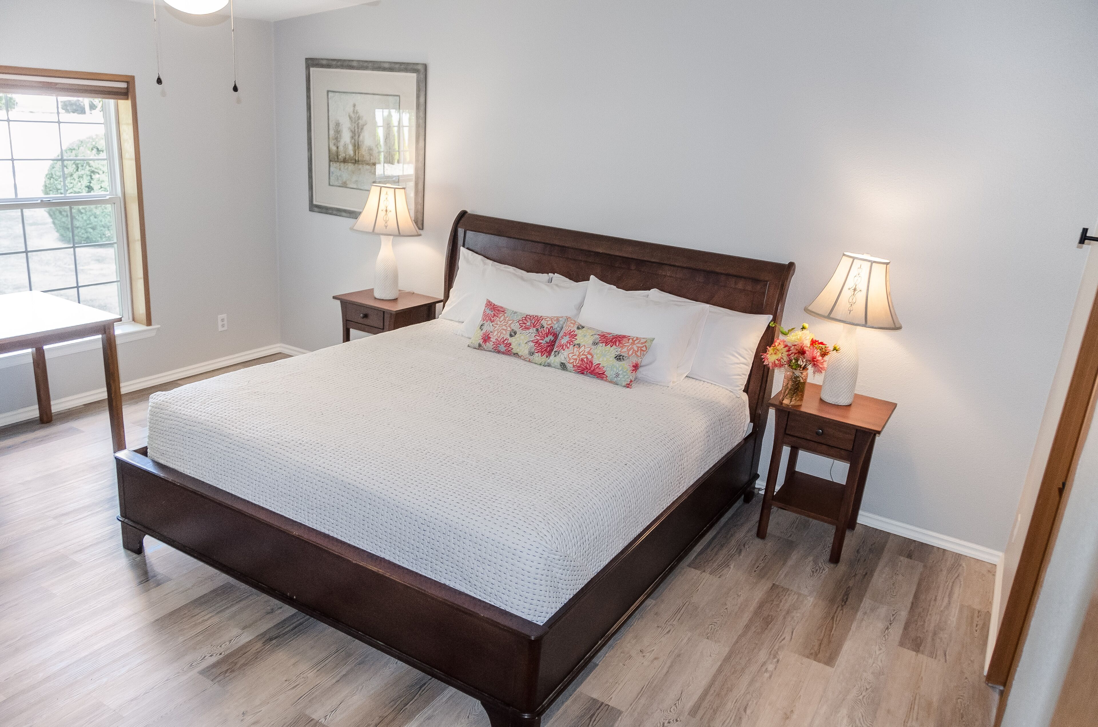 A neatly made bed with a wooden headboard, two bedside tables with lamps, and a window with a view of greenery.