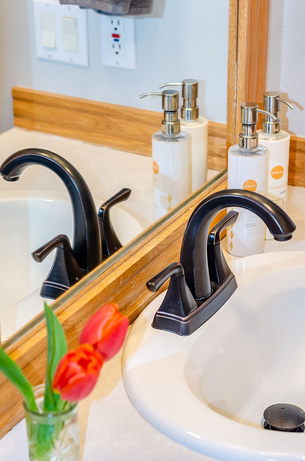 Ein Badezimmer mit einem weißen Waschbecken, schwarzen Wasserhähnen und einem Spiegel, der den Raum reflektiert. Auf der Ablage liegen zwei Lotionflaschen.