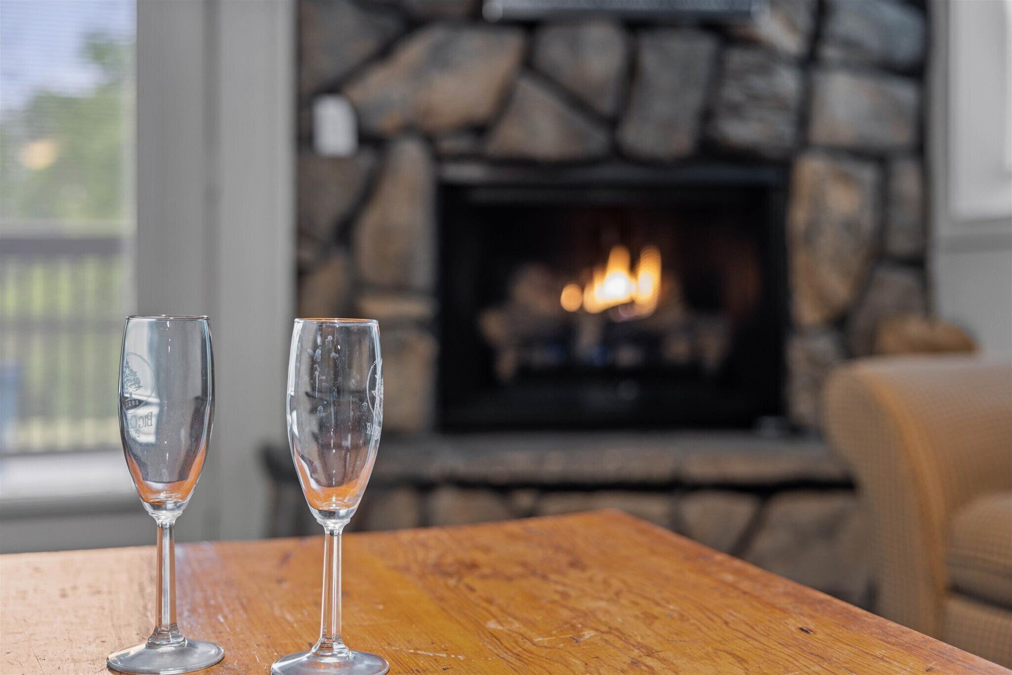Two empty wine glasses on a wooden table with a stone fireplace in the background.