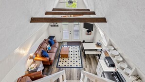 Ein Loft-Wohnbereich mit hoher Decke, Holzbalken, einer Sofagarnitur, einem Couchtisch, einem Fernseher und einer Küchenzeile mit Geschirr.