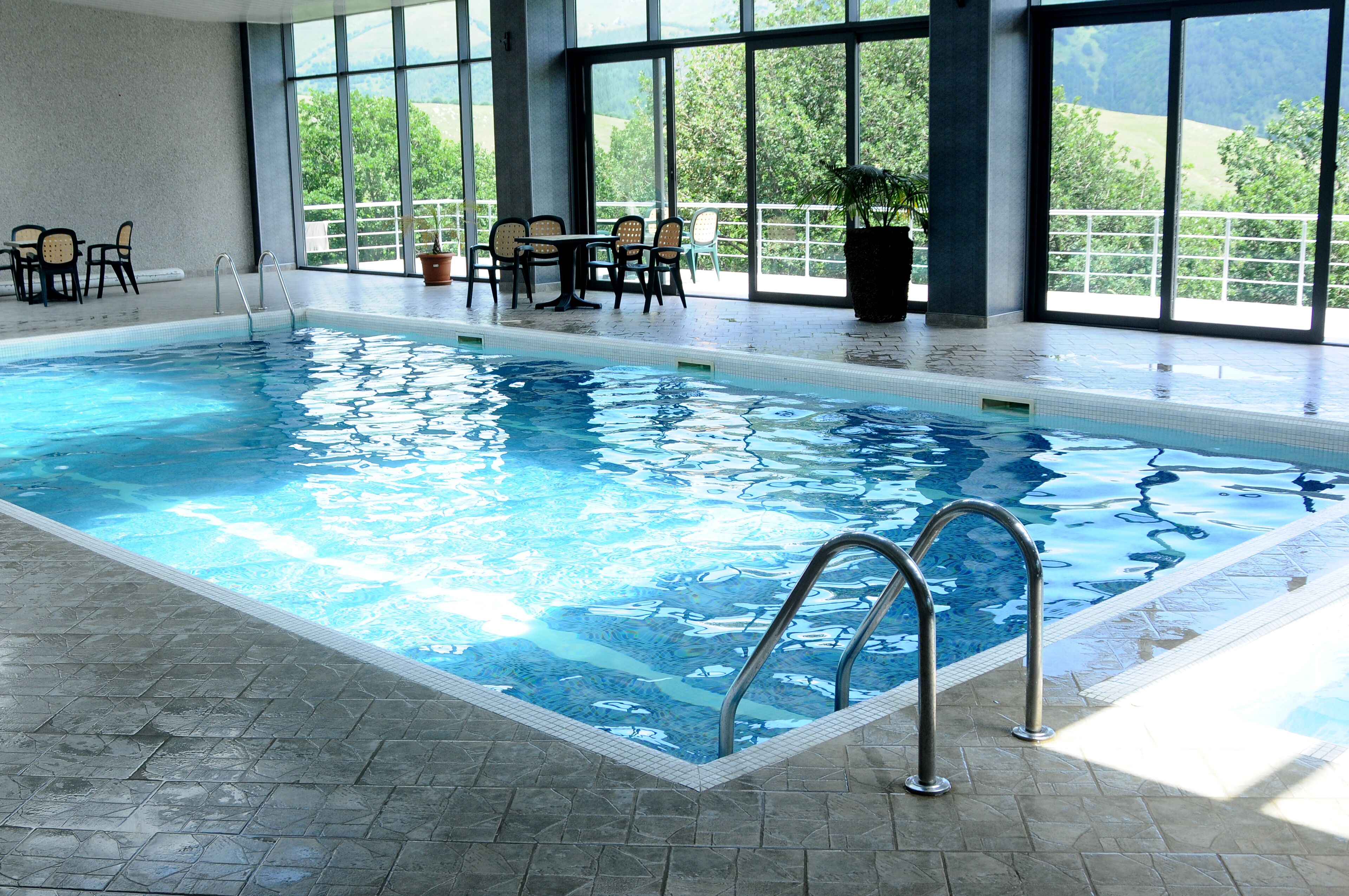 An indoor swimming pool with clear blue water, a tiled floor, and a seating area with chairs and tables.