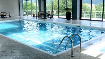 An indoor swimming pool with clear blue water, a tiled floor, and a seating area with chairs and tables.