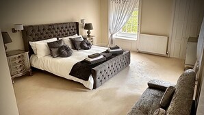Une chambre avec un grand lit, une table de chevet, une lampe, une fenêtre avec des rideaux et un banc.
