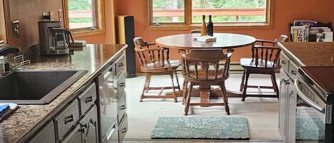 A kitchen with dark cabinets, a granite countertop, a dining area with a wooden table and chairs, and a large window with a view of trees.