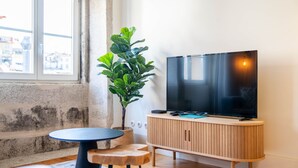 Een woonkamer met een televisie op een houten kast, een klein rond tafeltje en een grote groene plant.