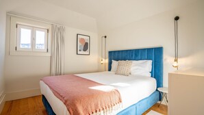 Een slaapkamer met een groot bed, een raam met gordijnen, een lijst met een afbeelding aan de muur, en een nachtkastje met een lamp.