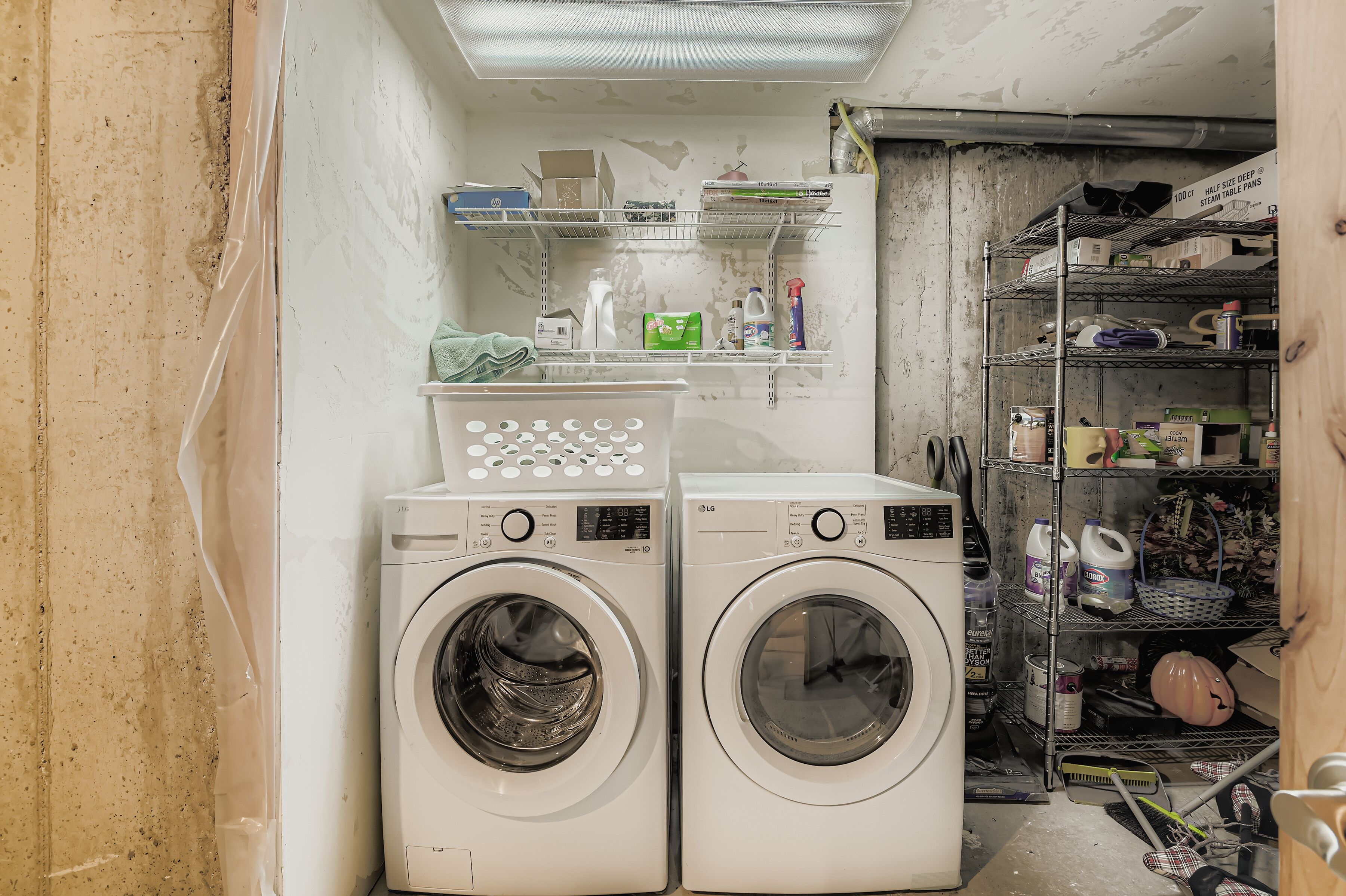 A laundry room with two front-loading washing machines, shelves with cleaning supplies, and a metal rack with various items.
