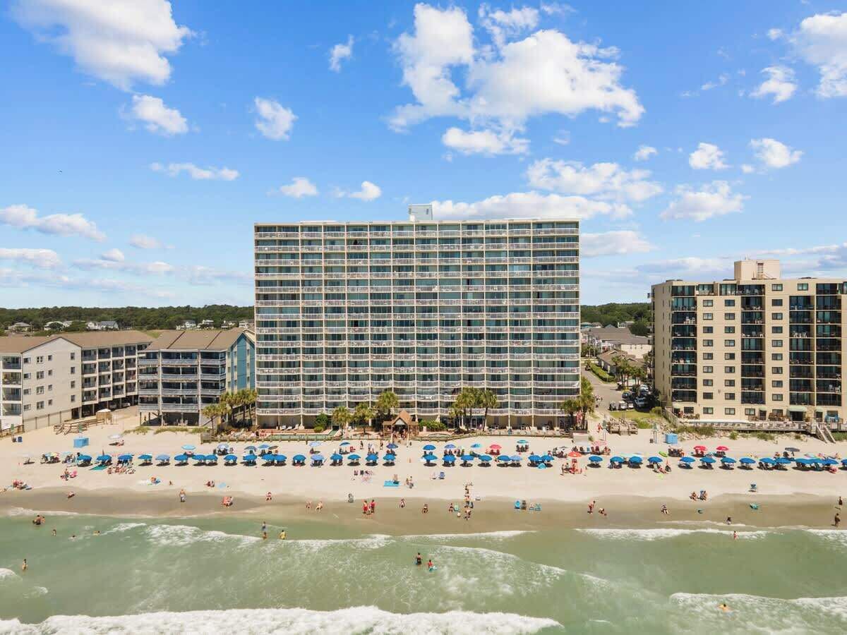 Een hotel aan het strand met meerdere verdiepingen, een strand met parasols en een heldere lucht.
