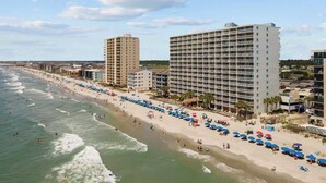 Un hôtel situé en bord de mer, avec plusieurs étages, une plage de sable et des parasols.