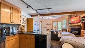 A compact, modern kitchen with wooden cabinets, a dishwasher, and a coffee maker. There is a dining area with a table and chairs, and a cozy living space with a fireplace and a bed. - Mountain Escape and Adventure at Winter Park (Winter Park)