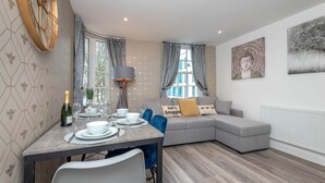 Ein modernes Wohnzimmer mit einem grauen Sofa, einem Esstisch mit Tellern und Gläsern sowie einem großen Fenster mit Vorhängen.