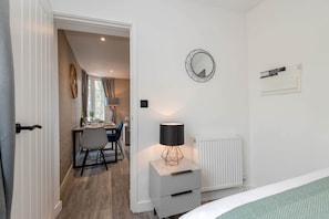 A bedroom with a bed, a nightstand with a lamp, a radiator, and a wall-mounted mirror. - The Honeycomb Residence by Fortuna Property Portfolio (Cheltenham)