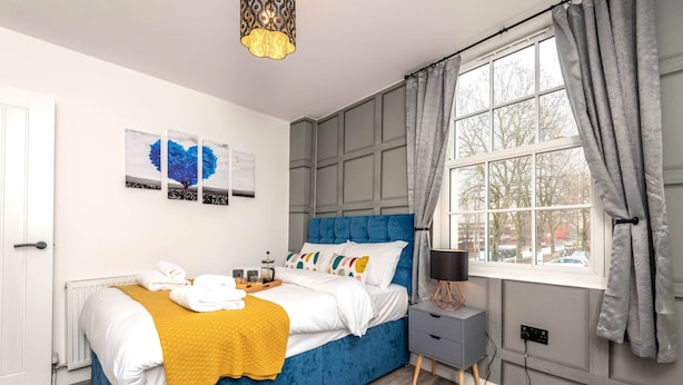 Ein modernes Schlafzimmer mit einem blauen Bett, weißer Bettwäsche, einer senfgelben Tagesdecke und einem Nachttisch mit einer Lampe.