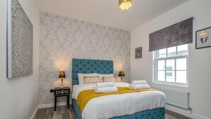 A bedroom with a tufted blue headboard, a white and yellow bedspread, two bedside lamps, and a large window with a view of the outside. - The Cotswold Woodland by Fortuna Property Portfolio (Cheltenham)