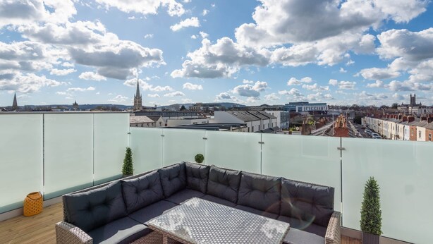 Eine Dachterrasse mit einer Sitzgelegenheit mit Kissen, einem kleinen Tisch und Blick auf die Stadtlandschaft, einschließlich eines Kirchturms.