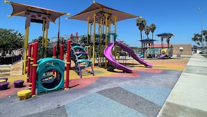 A playground with slides and climbing structures, surrounded by palm trees and a clear sky.