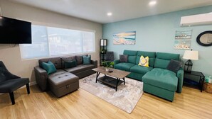 A modern living room with a brown sectional sofa, a green sofa, and a wooden coffee table. There is a TV mounted on the wall and a window providing a view of the outside.