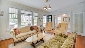 Un salón amplio con un sofá color beige, un sofá floral y un sillón a juego. Hay ventanas grandes con persianas, una araña y una puerta con paneles de vidrio.