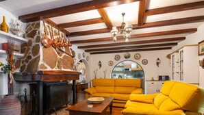 A living room with a stone fireplace, yellow sofa, wooden beams, and a kitchen visible through an archway.