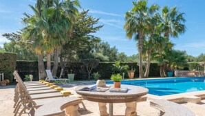 A poolside area with a circular stone table, lounge chairs, and a swimming pool surrounded by palm trees.