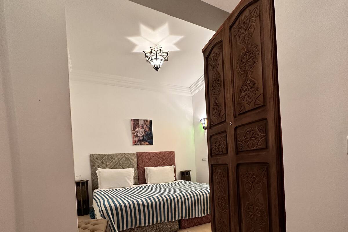 A bedroom with a bed, a nightstand, a patterned rug, a wooden wardrobe, and a ceiling light fixture.
