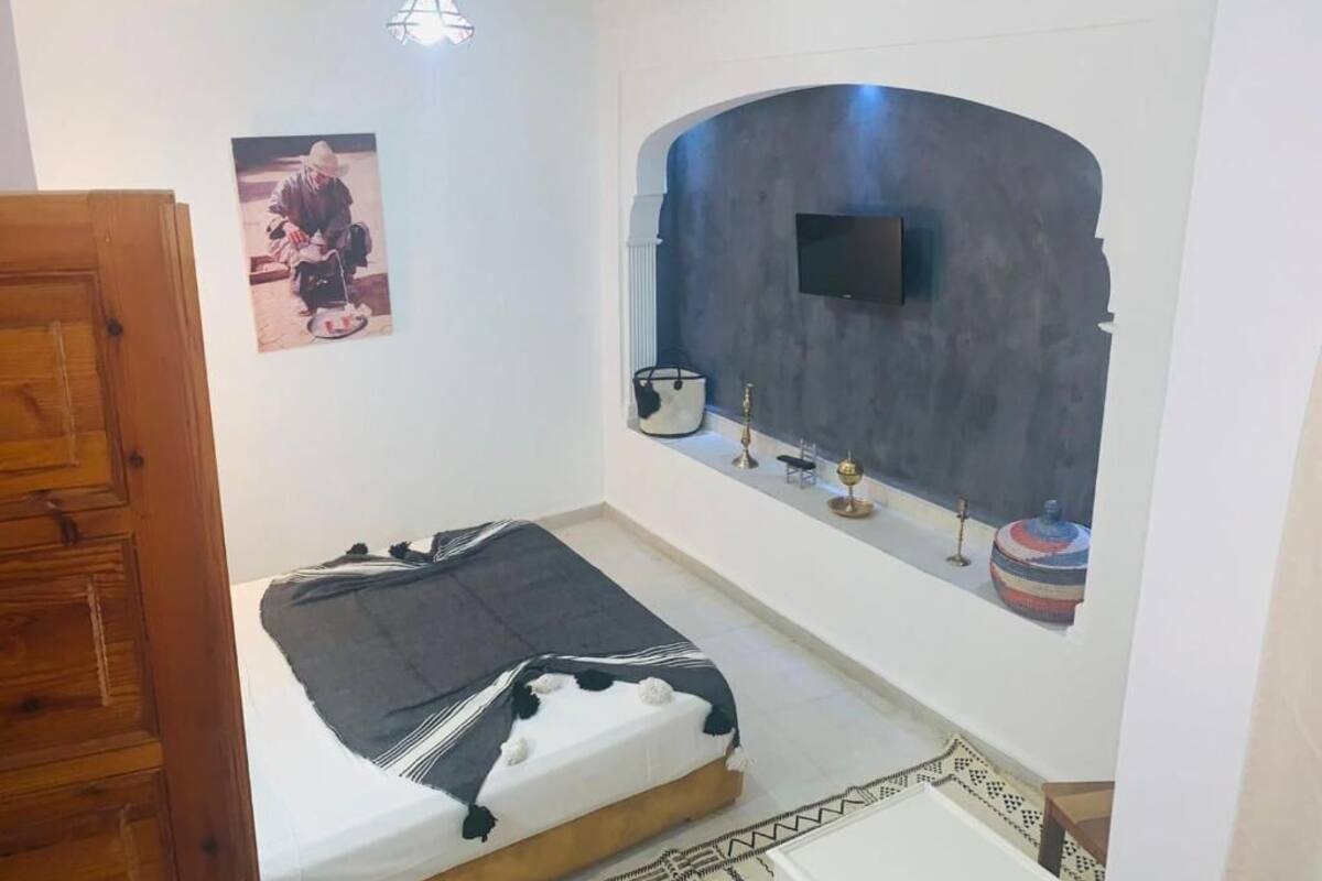 A bedroom with a bed, a television mounted on a curved wall, a wooden door, and a small shelf with decorative items.
