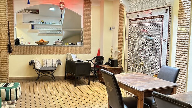 A dining area with a patterned floor, a table with chairs, a kitchen in the background, and a decorative wall. - Riad Les Miroires (Marrakech)